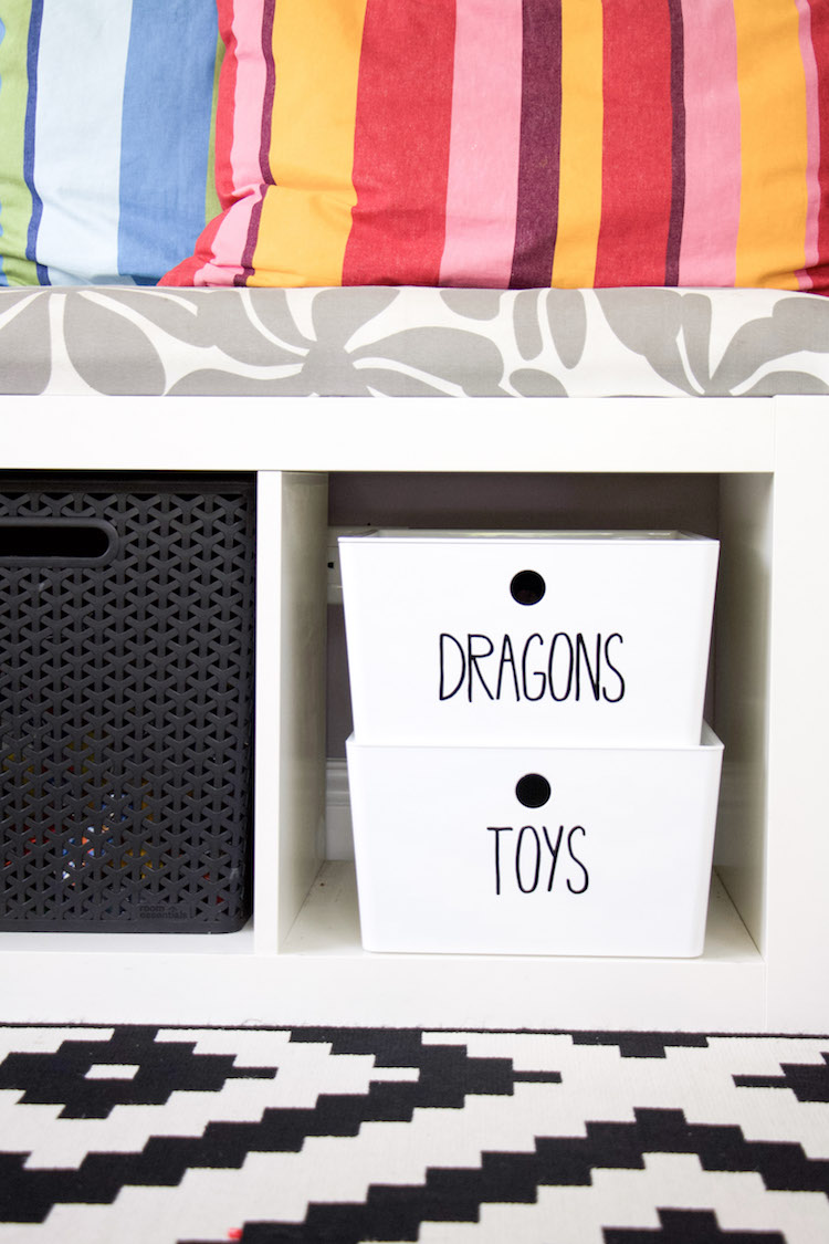 bench with cushion and colorful pillows. under bench two white bins with black labels that say dragons and toys to demonstrate toy storage of action figures