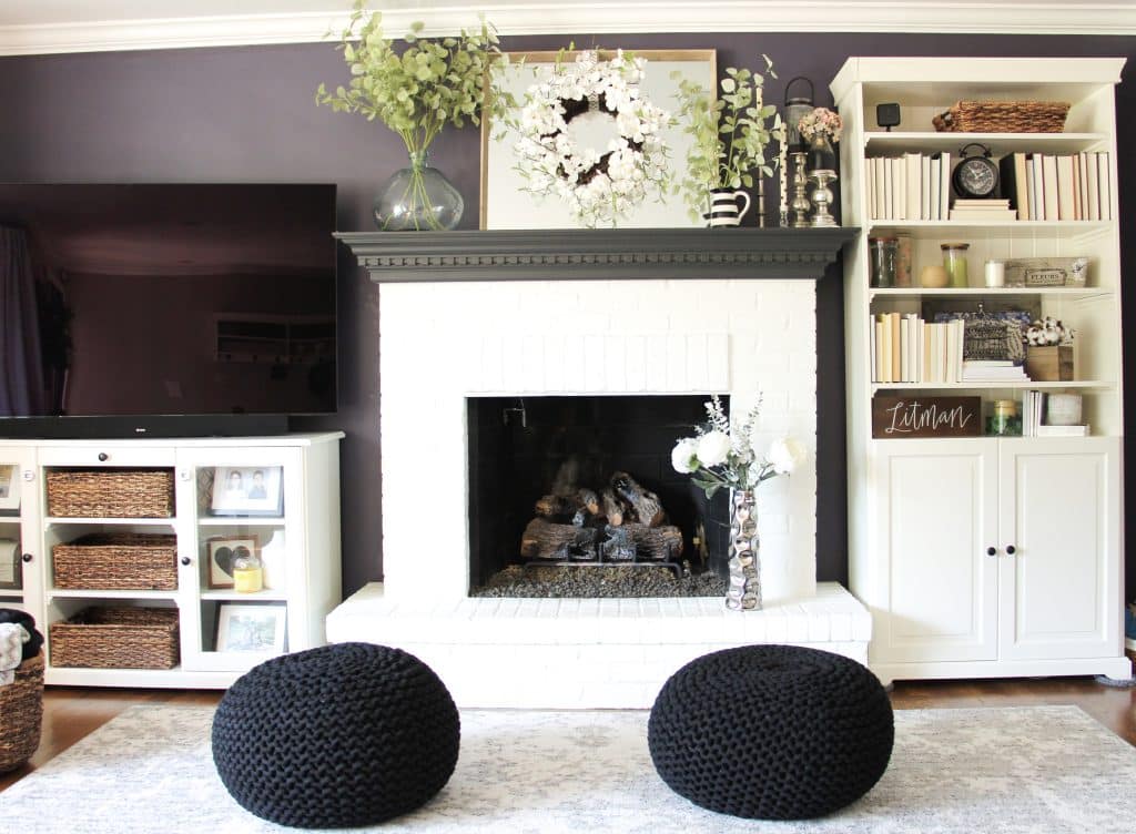 tv on stand next to white fireplace with a bookshelf to the right and two black poufs in front of the fireplace